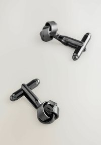 Juodos metalinės kaklaraiščio segtukai, turintys susuktos mazgo dizainą, su lygiu paviršiumi, apvaliais galais ir blizgiu tekstūra.