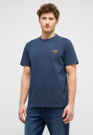 Man with curly hair wearing navy blue Mustang Jeans Co. t-shirt and blue jeans, standing against plain light gray background.