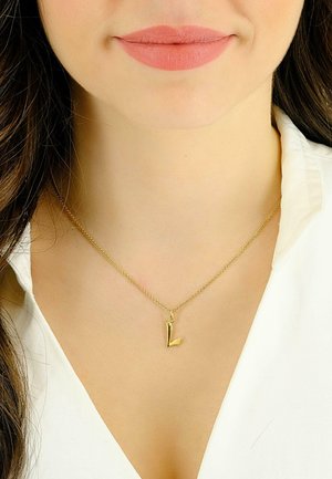 Collier pendentif en or avec un charm en forme de lettre 'l' minuscule sur une chaîne délicate, porté sur le décolleté d'une chemise blanche. Design simple et minimaliste.