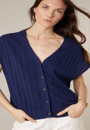 Femme portant un cardigan tricoté bleu marine à manches courtes avec boutons et col en V, mains dans les poches d'un pantalon blanc, fond neutre.