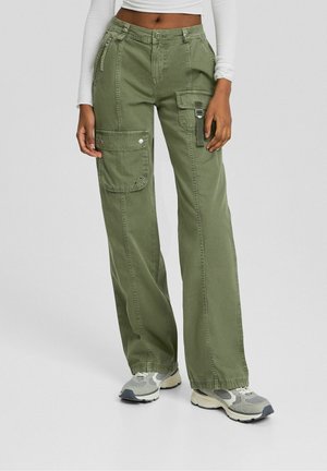 Frau trägt olivgrüne Cargo-Hose mit mehreren Taschen, kombiniert mit grauen und weißen Sneakern und einem weißen, kurzen Langarmtop.