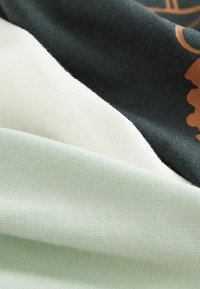 Textilní látka s třemi barvami: tmavě zelená, krémová a jemná máta. Textura je hladká s jemným leskem.
