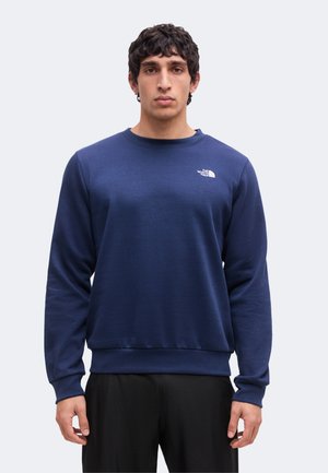 Homme aux cheveux courts et bouclés portant un sweat-shirt bleu marine uni avec le logo The North Face sur la poitrine et un pantalon noir.