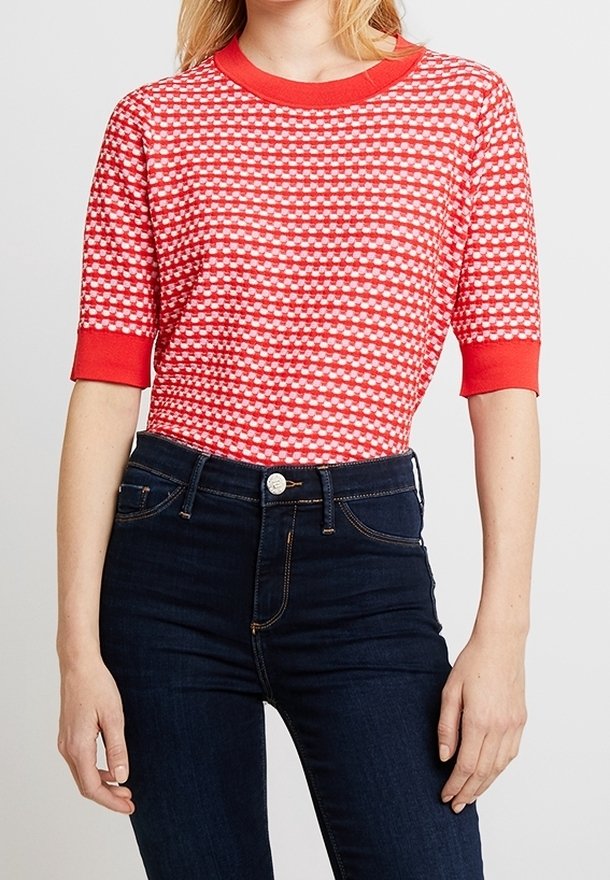 Une femme porte un pull à manches courtes à motifs rouges et blancs avec des poignets et un col rouges, associé à un jean bleu foncé taille haute.