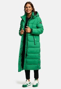 Manteau matelassé long et vert avec capuche, présentant un design matelassé, des accents noirs et des fermetures à boutons-pression. Porté sur un pantalon noir et des baskets.