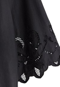 Svart tyg med dekorativ laserutskuren blommig broderi och scallopade kanter. Designen har intrikata mönster och öppna nät-detaljer.