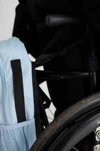 Light blue backpack with a black mesh pocket attached to a wheelchair, featuring black straps and a textured black wheel.