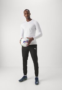 T-shirt de sport à manches longues blanche associé à un pantalon de jogging noir, arborant un logo Puma. Le mannequin tient un ballon de football coloré.