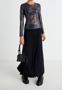 Žena v černém síťovaném topu s dlouhým rukávem, černé maxi sukni, držící malou černou kabelku a obutá v černých robustních botách na bílém pozadí.