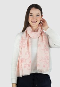 Lichtroze sjaal met ingewikkeld bloemenpatroon in donkerder roze, gemaakt van zachte stof, gedragen over een witte gestructureerde trui, met franjes aan de randen.