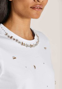 Witte T-shirt met een ronde halslijn, versierd met sprankelende heldere en gouden decoratieve elementen. Gladde stof met decoratieve versieringen.