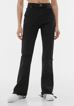 Personne portant un jean noir taille haute usé avec genoux et ourlets effilochés, associé à des baskets blanches sur un fond uni.
