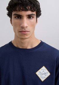Camiseta de algodón azul marino con cuello redondo, que presenta un parche blanco en forma de diamante con costuras amarillas y una marca de bar de cócteles.