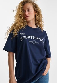 Mörkblå översized sport-t-shirt med korta ärmar, vit text som säger "SPORTSWEAR" och en rund logotyp. Mjuk tygtextur.