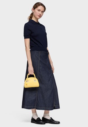 Mujer vestida con polo de manga corta azul marino, falda larga de mezclilla oscura, mocasines negros con calcetines claros, sosteniendo un pequeño bolso amarillo al lado.