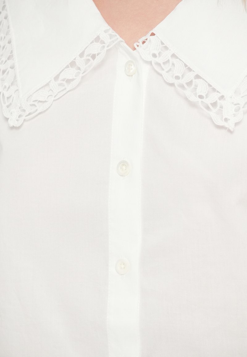 Weiße Bluse mit großem Spitzenkragen, Knopfleiste und glatter Stofftextur. Der Kragen hat ein filigranes Spitzenmuster.