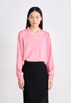Roze blouse met lange mouwen en een V-hals, voorzien van gather manchet, gecombineerd met een geribbelde zwarte rok. De stof lijkt lichtgewicht en glad.