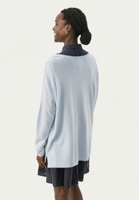 Hellblauer Pullover, gerippter Ausschnitt, lockere Passform, lange Ärmel, seitliche Schlitze. Wird über einem dunklen Kleid getragen, Modell sieht von der Kamera weg.