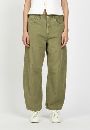 Person trägt weite olivgrüne Hose, weiße Sneaker und ein bauchfreies weißes Oberteil und steht vor einem schlichten, hellen Hintergrund.