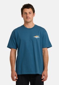 Mann mit kurzem braunem Haar und Schnurrbart, der ein blaues T-Shirt mit einem kleinen tropischen Inselmotiv auf der Brust trägt und vor einem weißen Hintergrund steht.