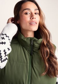 Green puffy vest with a high collar and front zipper, layered over a white sweater with a black patterned design. Smooth texture, casual fit.