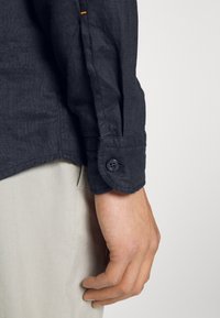 Main droite détendue, portant une chemise à manches longues foncée avec un poignet boutonné et un pantalon beige clair, sur un fond uni.