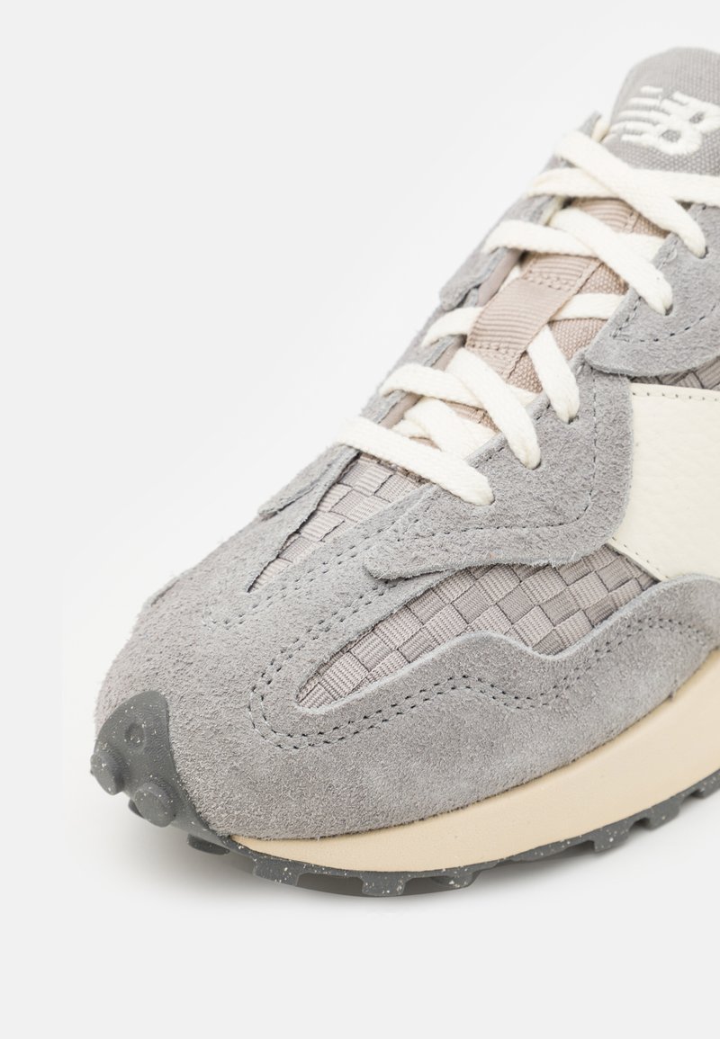 Zapatilla de ante gris con diseño de malla y acentos en color blanco roto. Incluye superposiciones texturizadas y una suela de goma con puntos de tracción.