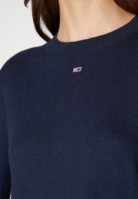 Gros plan d'une personne portant un pull bleu marine à col rond avec un petit logo rectangulaire rouge, blanc et bleu centré sous l'encolure.