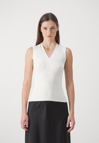 Top bianco senza maniche in tessuto a costine con scollatura a V e dettagli delle cuciture sottili, abbinato a una gonna nera plissettata.