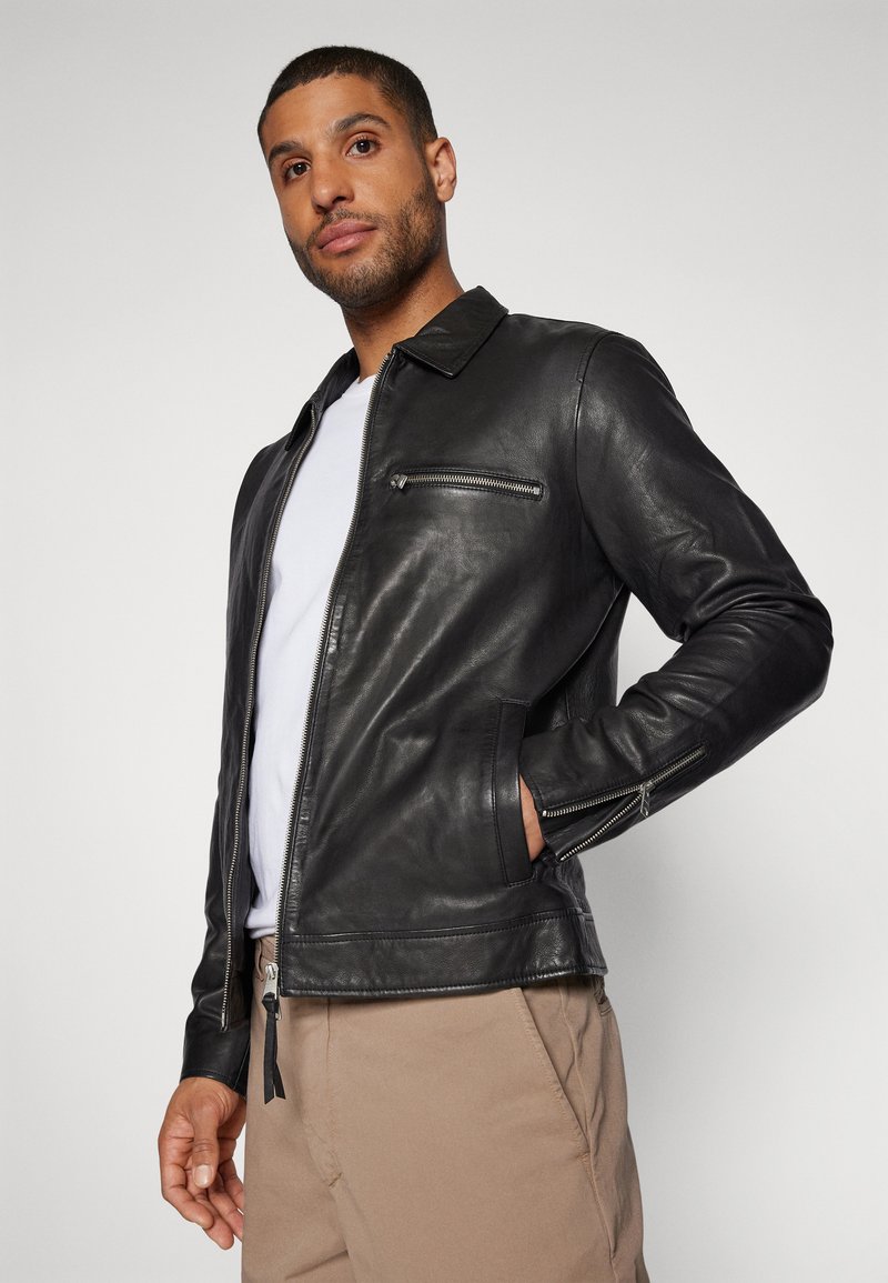 Mann in schwarzer Lederjacke über weißem Hemd und beigen Hosen, der mit einer Hand in der Tasche vor einem schlichten Hintergrund steht.