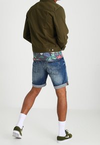Chaqueta de mezclilla verde oliva, camiseta de flores, pantalones cortos de mezclilla azules, calcetines blancos y zapatillas verdes. Vista trasera con los puños enrollados.