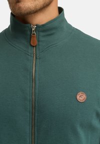 Mand med en teal ribbet zip-up jakke med en brun læder lynlås og en rund brun logo patch på brystet.