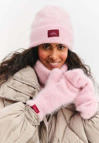 Różowy dziergany czapka, szalik i rękawiczki wykonane z miękkiego, puszystego materiału. Beżowa puchowa kurtka z widocznymi elementami zapięć na rękawie.