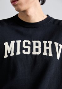 Mustas kootud särk, millel on kreemja plokktekstiga "MISBHV", ribakudumisega kaela- ja õlavarredetailid.