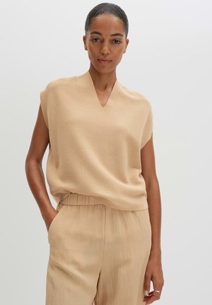 Femme aux cheveux foncés portant un haut beige sans manches à col en V et un pantalon large assorti, debout avec une main dans la poche sur un fond uni.
