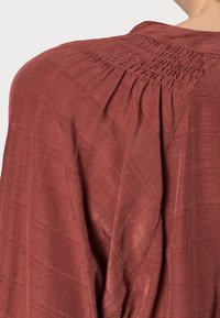 Blusa color ruggine con tessuto texturizzato e intrecciato. Presenta dettagli arricciati sul retro e una vestibilità rilassata con una superficie leggermente decorata.
