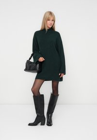 Tumši zaļš pārāk liels adīts kleitas svārks ar garām piedurknēm, kopā ar melniem augstiem zābakiem un mazu melnu rokassomu ar teksturētu apdari.