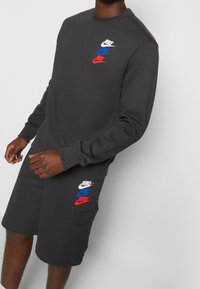 Tmavě šedá mikina a odpovídající kraťasy. Mikina je ozdobena třemi barevnými logy Nike. Kraťasy mají boční kapsy a detaily s logem.