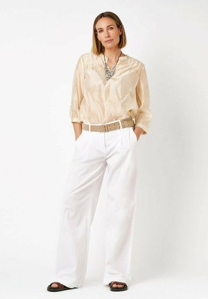 Frau steht mit den Händen in den Taschen, trägt eine beige Bluse, weite weiße Hosen, einen braunen Gürtel, schwarze Sandalen und eine auffällige Halskette vor weißem Hintergrund.