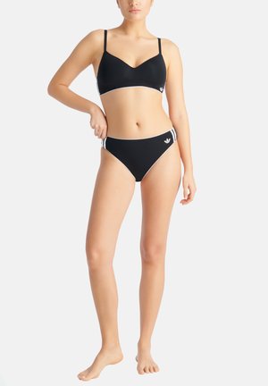Femme portant un soutien-gorge de sport noir assorti et un bas de bikini avec un liseré blanc et le logo Adidas, debout pieds nus sur fond blanc.