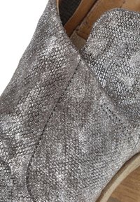 Zapato de textura plateada con un patrón de piel de reptil, con puntera redondeada y acabado en material suave, montado sobre una suela color marrón claro.