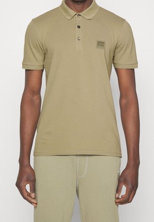 Homme portant un polo manches courtes vert olive clair avec deux boutons et un pantalon assorti, debout devant un fond uni.