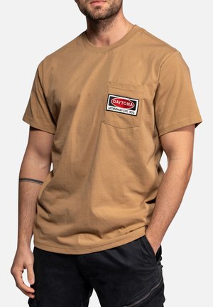 Homme portant un t-shirt beige à manches courtes avec un patch sur la poche poitrine indiquant "DAYTONA LEGENDARY EST. 1973" et un short noir avec une poche zippée.