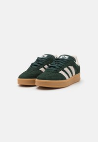 Green suede sneaker with white three-stripe design, cream accents at the heel, and a textured gum rubber sole. Laced front.