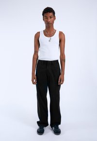 Pantalones negros de pierna ancha, top sin mangas blanco de canalé, collares de plata en capas, tatuajes visibles en los brazos, de pie contra un fondo blanco liso.