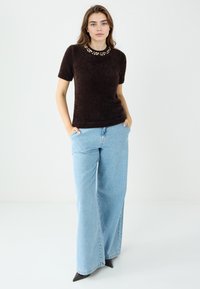 Brauner Kurzarmpullover mit strukturierter Oberfläche und verziertem Ausschnitt, kombiniert mit hoch taillierten hellblauen Jeans mit weitem Bein und schwarzen Absätzen.