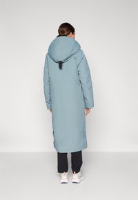 Cappotto lungo e blu chiaro imbottito con un grande cappuccio, cinturino regolabile sul retro, fessure laterali e una silhouette elegante, abbinato a pantaloni neri e sneakers.