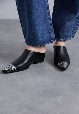Mule in pelle nera con punta affilata, dettagli in argento con borchie sul fronte, tacco basso a blocco, abbinate a jeans denim blu a gamba larga.
