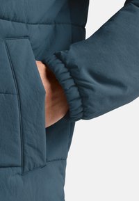 Giacca a vento teal con finitura testurizzata, dotata di tasca laterale e polsini elasticizzati. Le cuciture visibili aggiungono un dettaglio strutturale.