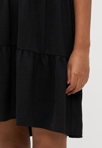 Svart klänning med en strukturerad tyg, med en volang nederkant i lager. Synlig hand som håller tyget vid botten.